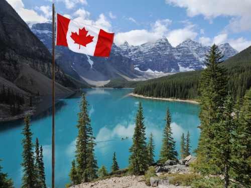 Lago  rodeado de vegetación con grandes montañas nevadas al fondo y la bandera de Canadá ondeando en primer plano