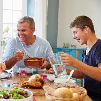 Hombre de mediana edad y chico adolescente riendo, sentados a la mesa y sirviéndose comida.