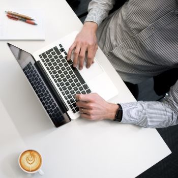Vista aérea de un hombre trabajando en un ordenador en una mesa blanca y un café con leche al lado