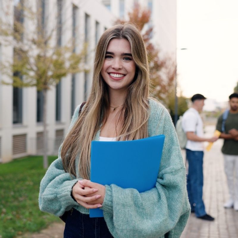 chica rubia sujetando una carpeta frente a una universidad en el extranjero
