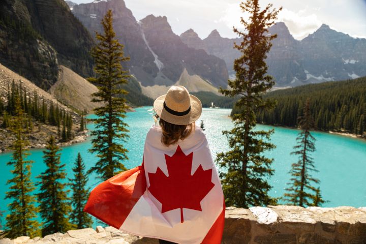 Chica envuelta con la bandera de Canadá contemplando un lago rodeado de montañas y vegetación espctacular