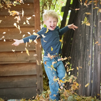 Niño rubio en un jardín jugando con hojas caídas mientras salta muy alegre.