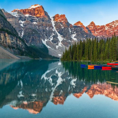 Fotografía de las Rocky Mountains reflejadas sobre un lago cristalino en Alberta, Canadá, con árboles verdes y canoas sobre el lago