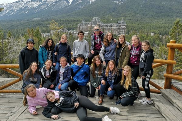Fotografía de un grupo de estudiantes de excursión, con las montañas nevadas al fondo, un monumento parecido a un castillo y un bosque frondoso alrededor
