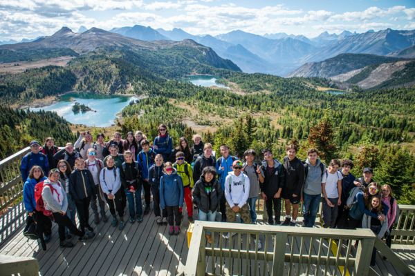 Fotografía de un grupo grande de estudiantes de excursión en un mirador de madera desde el que podemos apreciar las vistas de lo que parece una zona de parque natural con un bosque verde con lagos de color azul brillante en medio y montañas al fondo, en un tiempo soleado y cielo mayormente despejado