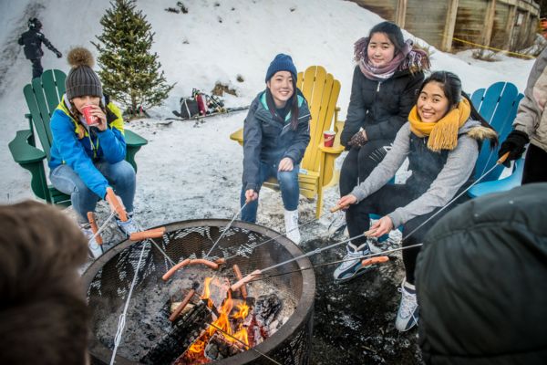 Fotografía de cuatro chicas alrededor de una hoguera sentadas tomando un chocolate caliente y asando salchichas en el fuego con palos, sonrientes y con los esquís puestos, de ve la nieve alrededor y una estructura tipo cabaña de madera detrás