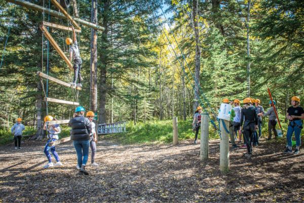 Fotografía de un grupo de estudiantes con cascos, dos trepando por una estructura tipo escalera hecha con tablas de madera y  cuerdas y otros esperando su turno, en un bosque soleado lleno de abetos