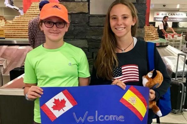 Fotografía de una niña canadiense junto a una española a la que ha ido a recoger al aeropuerto, las dos posan sonrientes sujetando un cartel de bienvenida hecho a mano con las banderas de Canadá y España