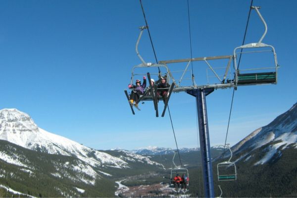 Fotografía de un telesilla a mucha altura, con las montañas nevadas de fondo y desde el que saludan estudiantes con los esquís puestos