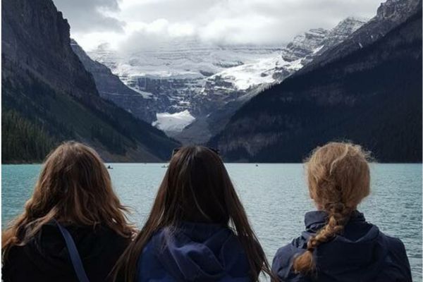 Fotografía de tres niñas de espaldas mirando al horizonte, donde se aprecia un lago azul y al fondo unas montañas cercanas que se abren y dejan ver más montañas nevadas y nubes blancas que dejan pasar unos rayos de sol entre nubes grises