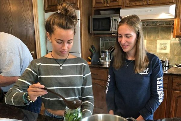 Fotografía en la que se ve a dos niñas cocinando, una con un cucharón en la mano y la otra mirándola sonriente, en una cocina con aire familiar y en lo que parece ser un aprendizaje en hacer un plato típico canadiense