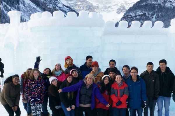 Fotografía de un grupo de estudiantes abrigados y muy contentos con su monitora, de excursión en las montañas nevadas con un castillo hecho de hielo y la parte superior curva hecha con nieve