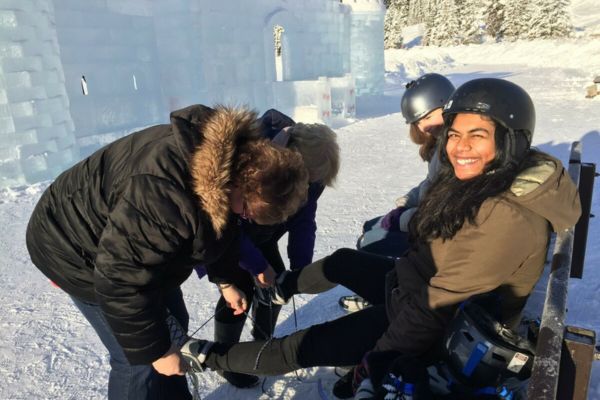 Fotografía de dos adultas atando los esquís a dos niñas, que miran a cámara y sonríen con cascos puestos y abetos nevados de fondo, así como un castillo de hielo de fondo y nieve en todo el suelo