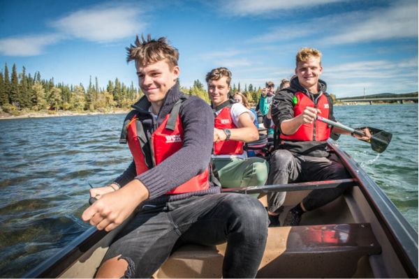 Fotografía de varios estudiantes en una canoa, tres chicos en primer plano sonrientes y remando en medio de un lago con árboles a lado izquierdo y tiempo soleado y cielo mayormente despejado