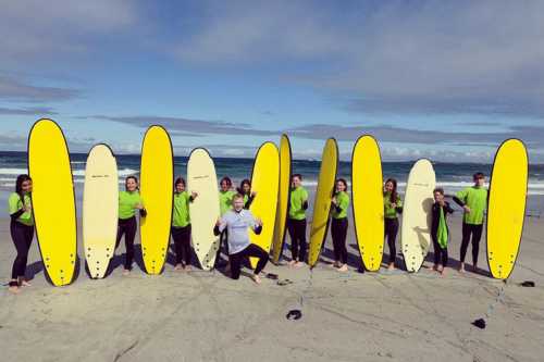 Chicos y chicas en fila mirando a la cámara en una playa irlandesa con tablas de surf amarillas al lado y el profesor delante agachado