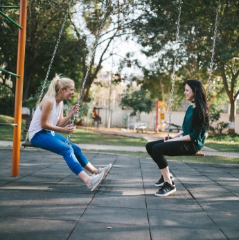 Dos chicas adolescentes subidas a un columpio uno frente a la otra conversando animadamente y sonriendo.