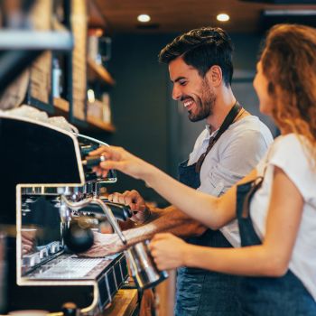 Barista realizando un café con expresión alegre, al lado tiene a una compañera que aparece de perfil  mirando hacía el realizando otro café