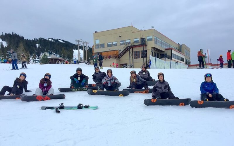 Grupo de alumnos con tablas de snowboard sentados en la nieve