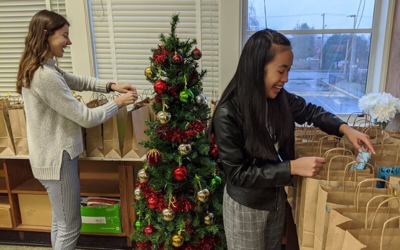 Dos chicas preparando bolsas para regalos de navidad con un pequeño árbol de navidad entre medio de las dos