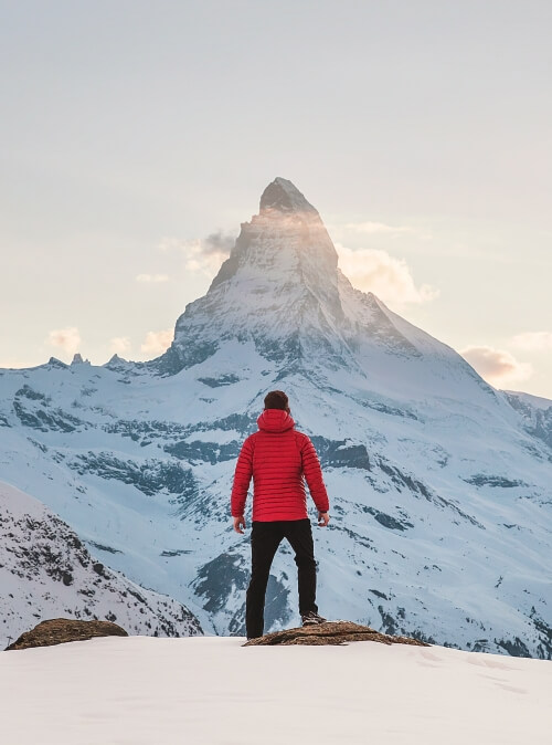 Chico con abrigo de montaña rojo, mirando a una montaña nevada en Suiza