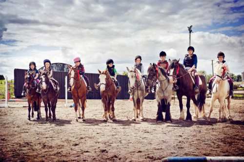 Niños y adolescentes con cascos de equitación en fila mirando a cámara subidos a ponis