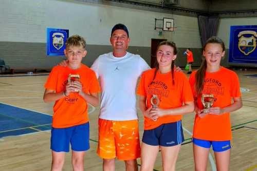 Tres alumnos, dos chicas y un chico,  vestidos con camisetas naranjas, junto con el entrenador de baloncesto en fila mirando a cámara agarrando un pequeño trofeo cada uno