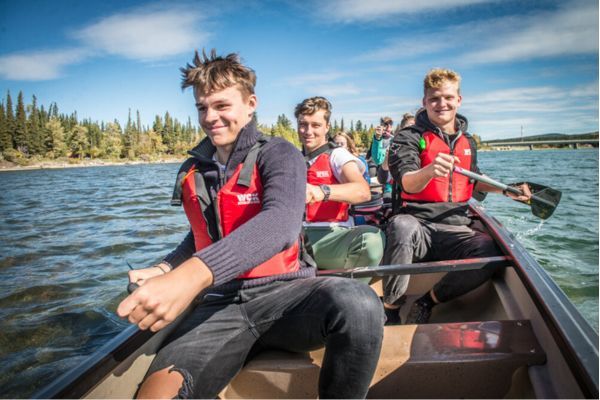 Fotografía de varios estudiantes en una canoa, tres chicos en primer plano sonrientes y remando en medio de un lago con árboles a lado izquierdo y tiempo soleado y cielo mayormente despejado
