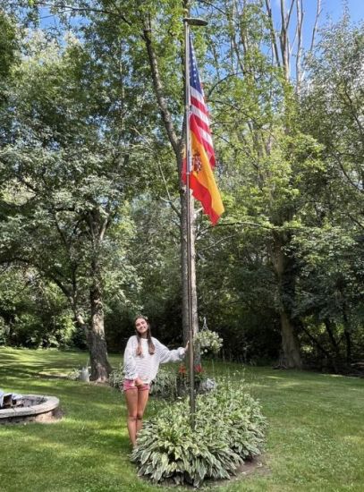 Alumna participante el programa J1 en Estados Unidos en el jardín de su casa anfitriona apoyada en un poste con una bandera Española y otra Americana 