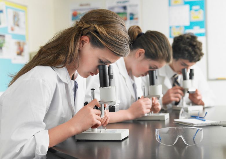 Alumnos adolescentes con bata blanca sentados de perfil mirando a través de un microscopio